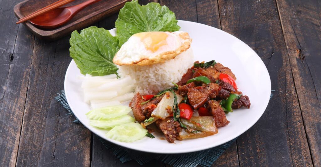 Appetizing Thai dish with stir-fried meat, rice, and fried egg served on rustic wooden table.